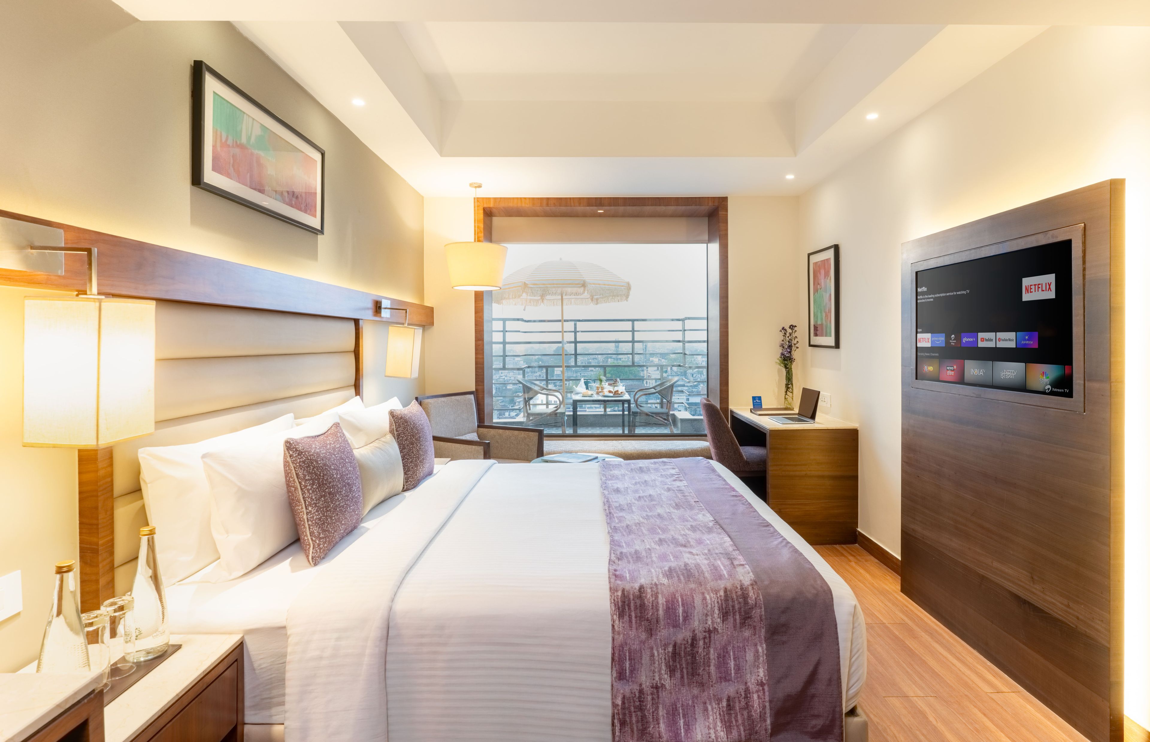 A hotel room with a bed, bedside tables, a desk with a laptop, a TV, and a balcony with a view.
