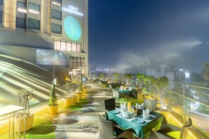 An outdoor dining area with tables and chairs, potted plants, and a building with illuminated signage. - Nirvana Sarovar Portico Gorakhpur (Gorakhpur)