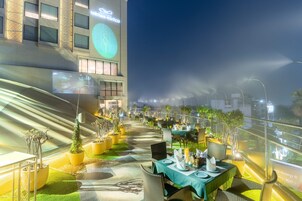 Un área de comedor al aire libre con mesas y sillas, plantas en macetas y un edificio con señalización iluminada.