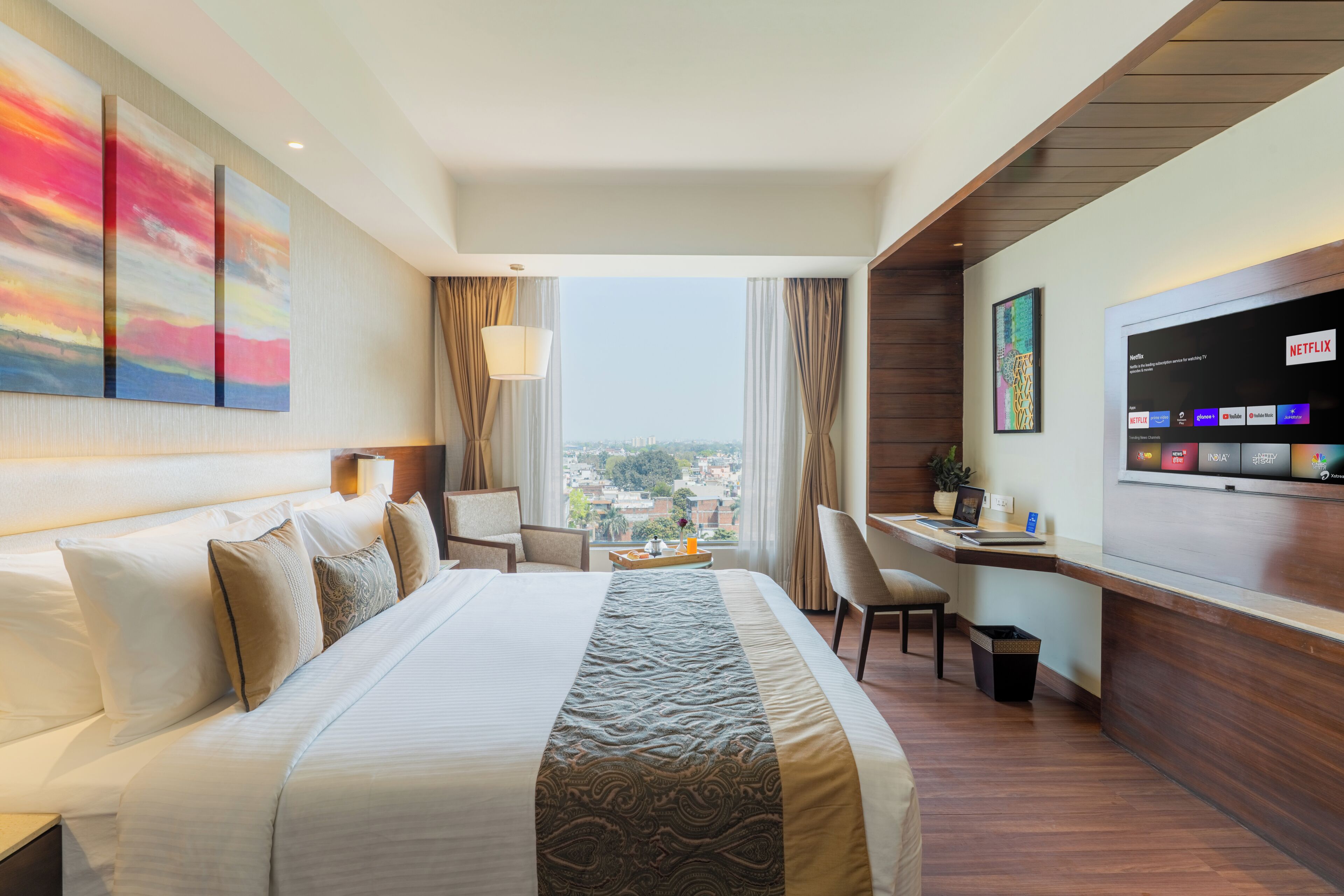 A hotel room with a large bed, a desk, a chair, a TV, and a view of the city.