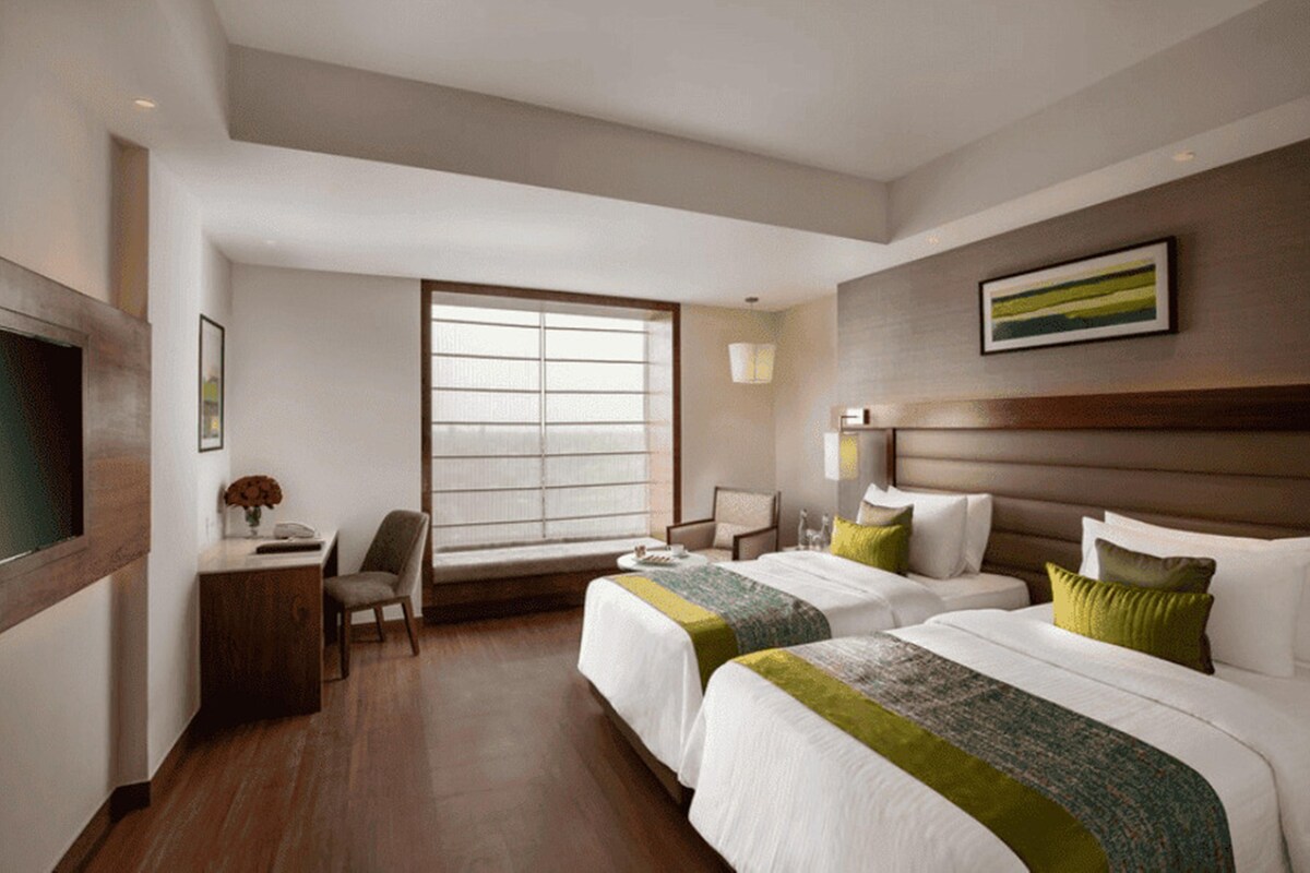 A hotel room with two beds, a desk, a chair, a window, and a framed picture on the wall.