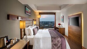 A hotel room with a large bed, a desk with a lamp, a TV, and a view of a cityscape. - Nirvana Sarovar Portico Gorakhpur (Gorakhpur)