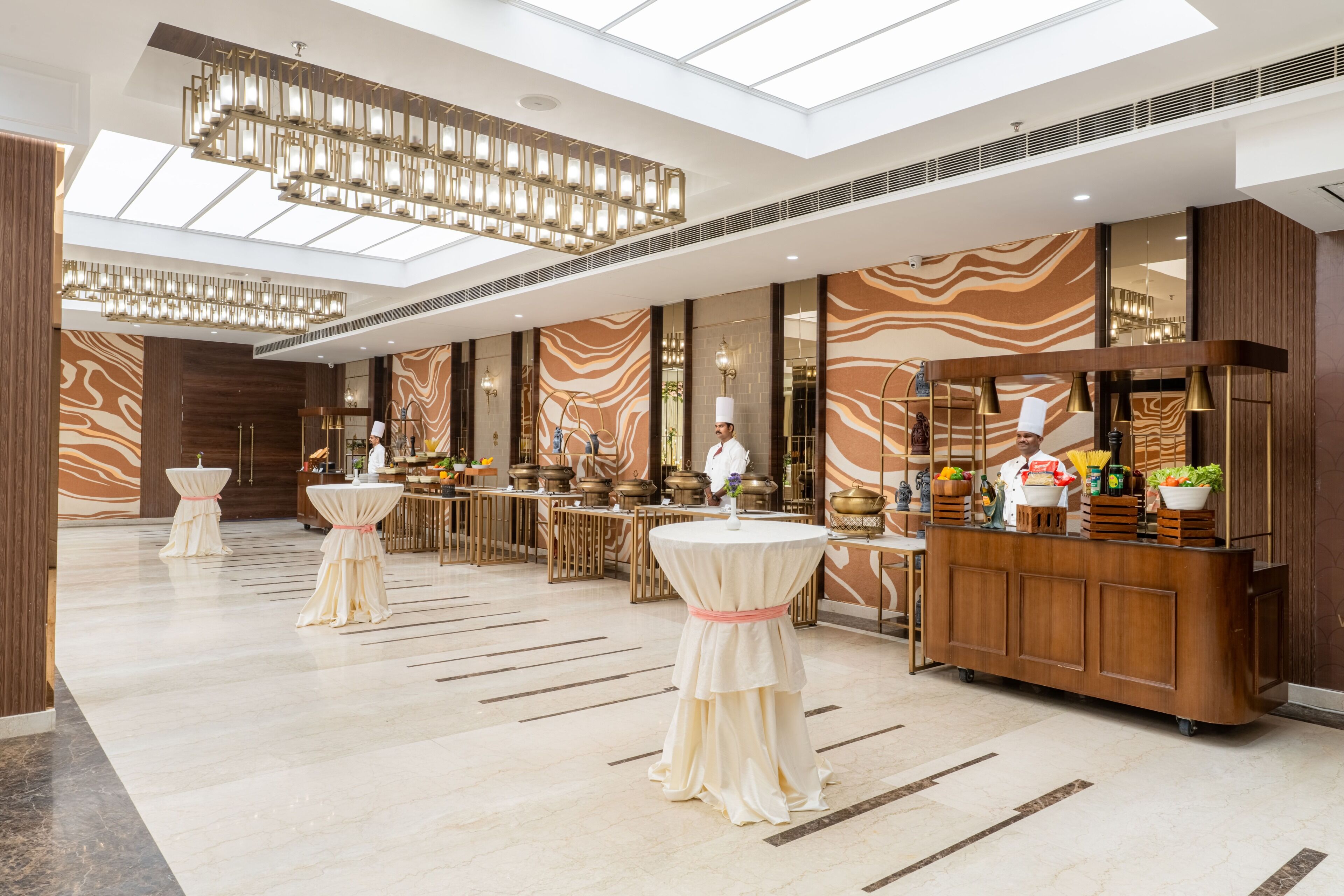 A modern hotel lobby with a high ceiling, chandeliers, a reception desk, and a bar area.