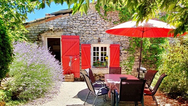 Un bâtiment en pierre avec un chemin de gravier menant à une table et des chaises placées sous un parasol rouge.