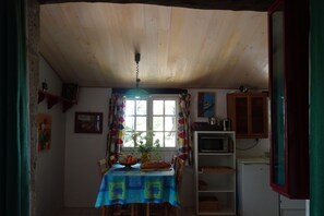 Ein Essbereich mit Tisch, der mit einer blauen Tischdecke bedeckt ist, Holzsesseln, einer Hängeleuchte, einem Fenster mit Vorhängen sowie einer Küchenzeile inklusive Mikrowelle und Schränken.