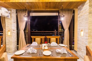 A dining table set for four with a city view, wooden beams, stone walls, and a balcony with curtains.