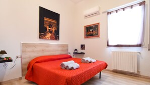 Une chambre à coucher comprenant un lit, une table de chevet, un climatiseur fixé au mur, une fenêtre avec des rideaux et des œuvres d’art accrochées au mur.