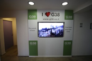A hallway with a wall-mounted TV displaying a cityscape, flanked by green banners with the G38 logo and the text "I ♥ G38". - G38 Rental Apartment Building (Haifa)