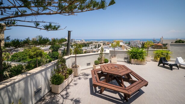 Une terrasse sur le toit, dotée d’une table et de chaises hexagonales, entourée de plantes en pot et offrant une vue sur un paysage urbain avec ses bâtiments et la mer.