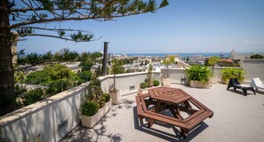 Una terraza en la azotea con una mesa y sillas hexagonales, rodeada de plantas en macetas y vistas a un paisaje urbano con edificios y el mar.