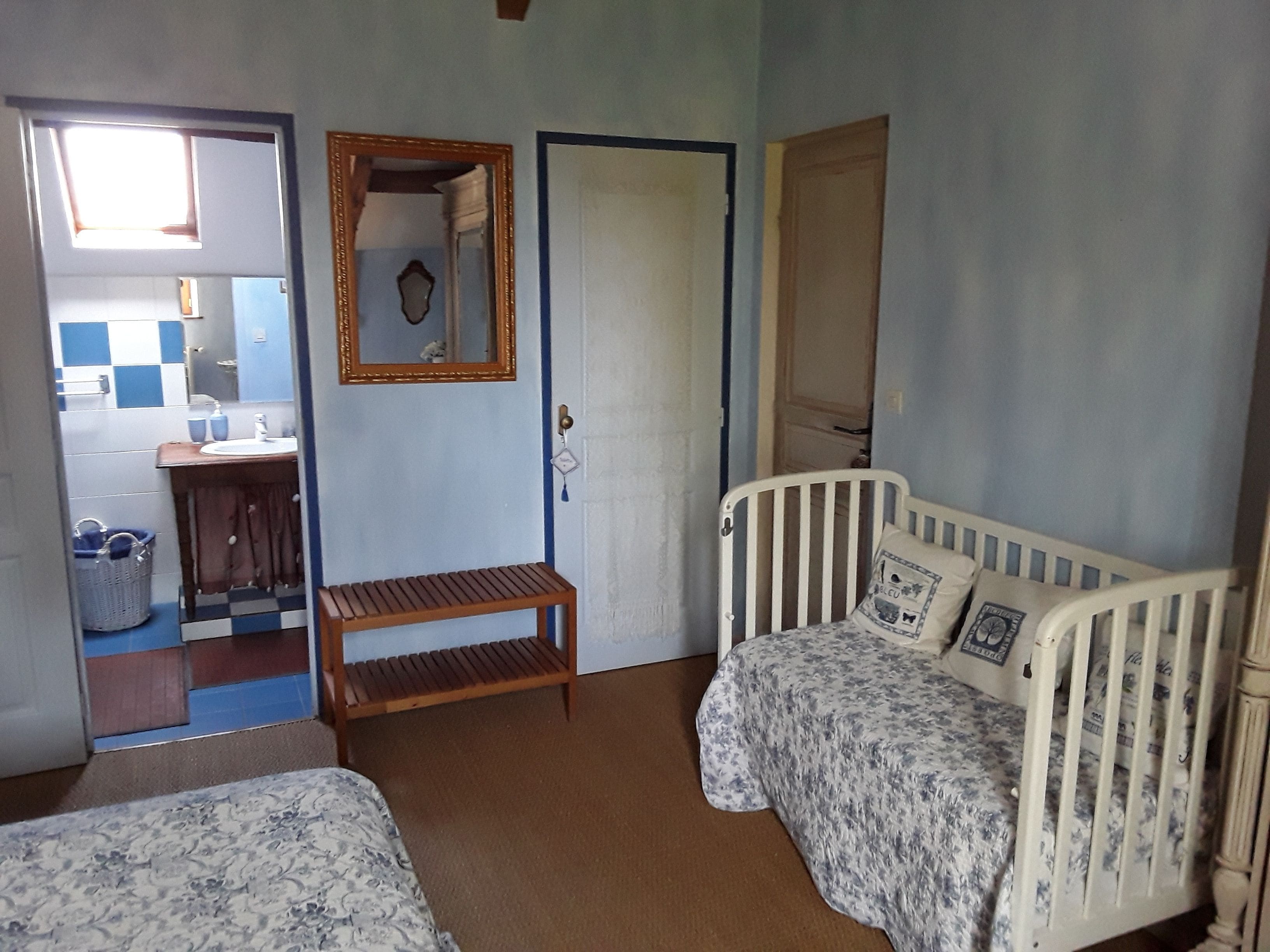 Une chambre avec deux lits, un banc en bois, un miroir et une porte donnant sur une salle de bain.