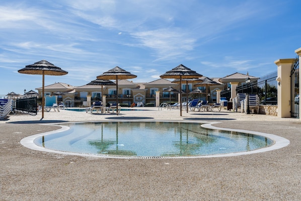 Un espace piscine agrémenté de chaises longues et de parasols, entouré d’un bâtiment doté d’un toit en tuiles.