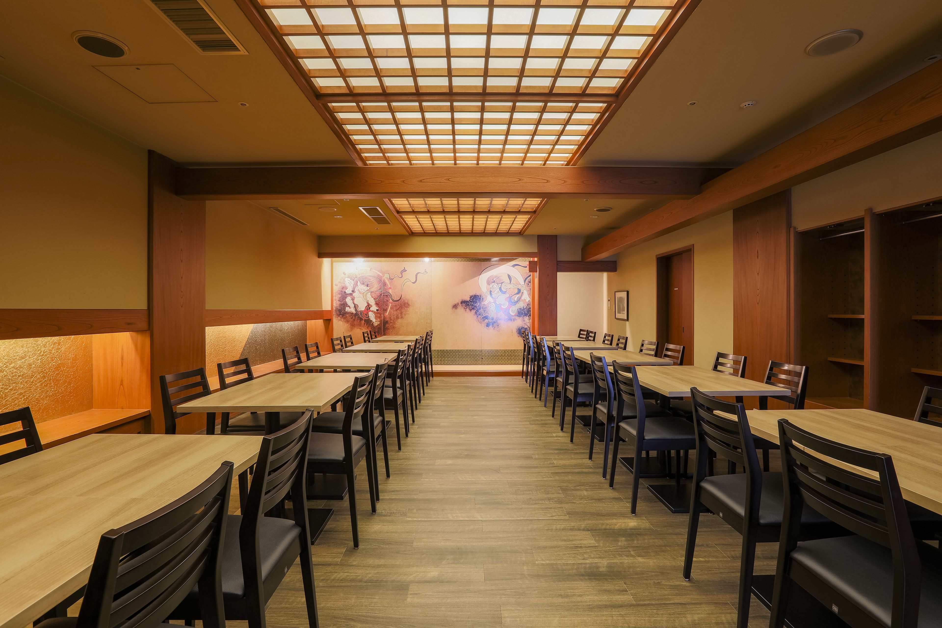 A restaurant with long wooden tables and black chairs, a tiled ceiling, and a mural on the back wall.