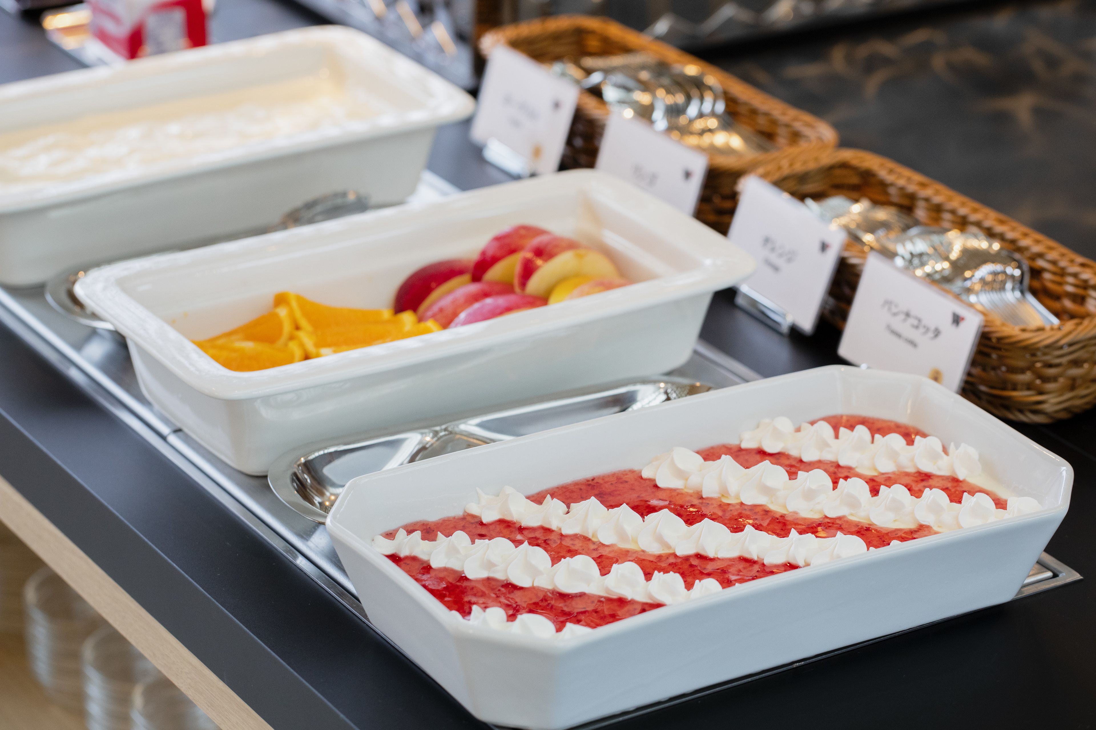 A buffet with various food items in white trays, including fruits and a dessert with whipped cream.