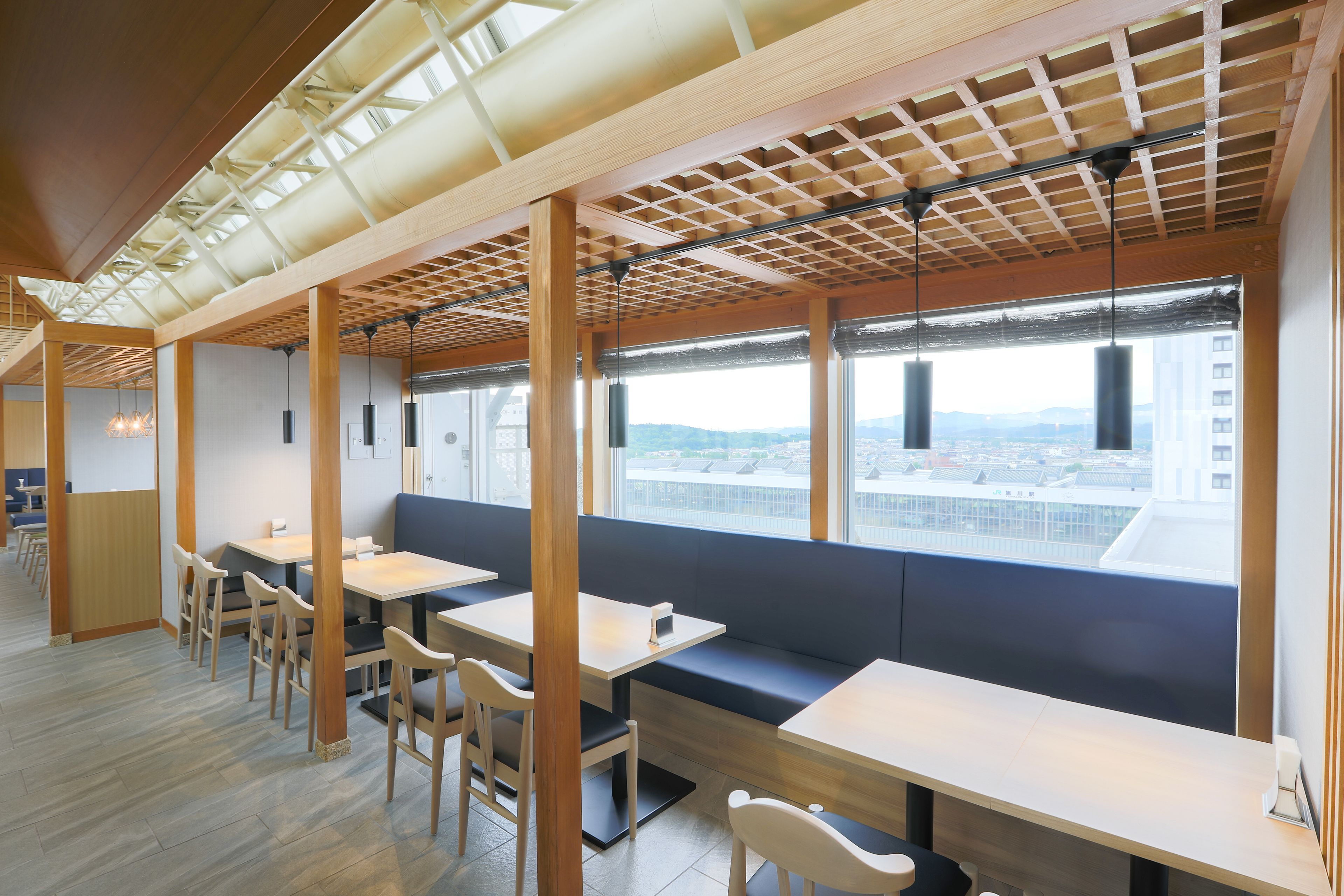 Un restaurante moderno con mesas y bancos de madera, grandes ventanales con vistas a la ciudad y un techo alto con vigas a la vista.