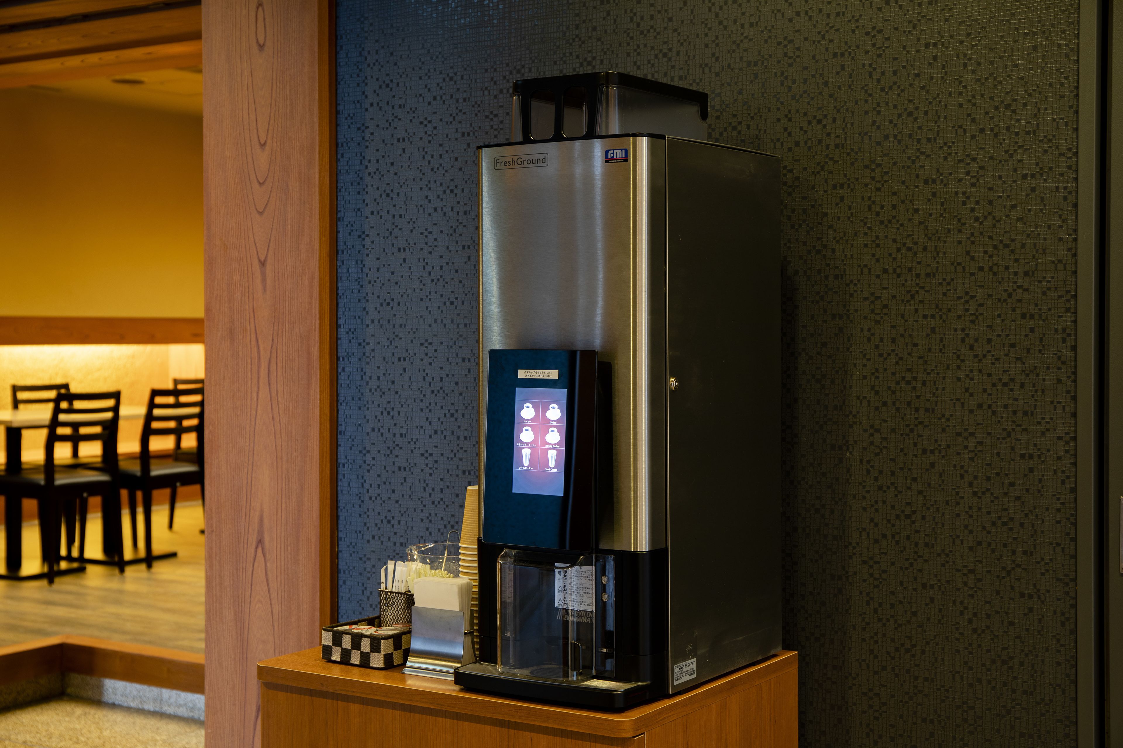 Une machine à café dotée d’une interface numérique, posée sur un comptoir en bois dans une pièce dont le mur est décoré d’un motif et où l’on aperçoit, au fond, un espace dédié à la salle à manger.