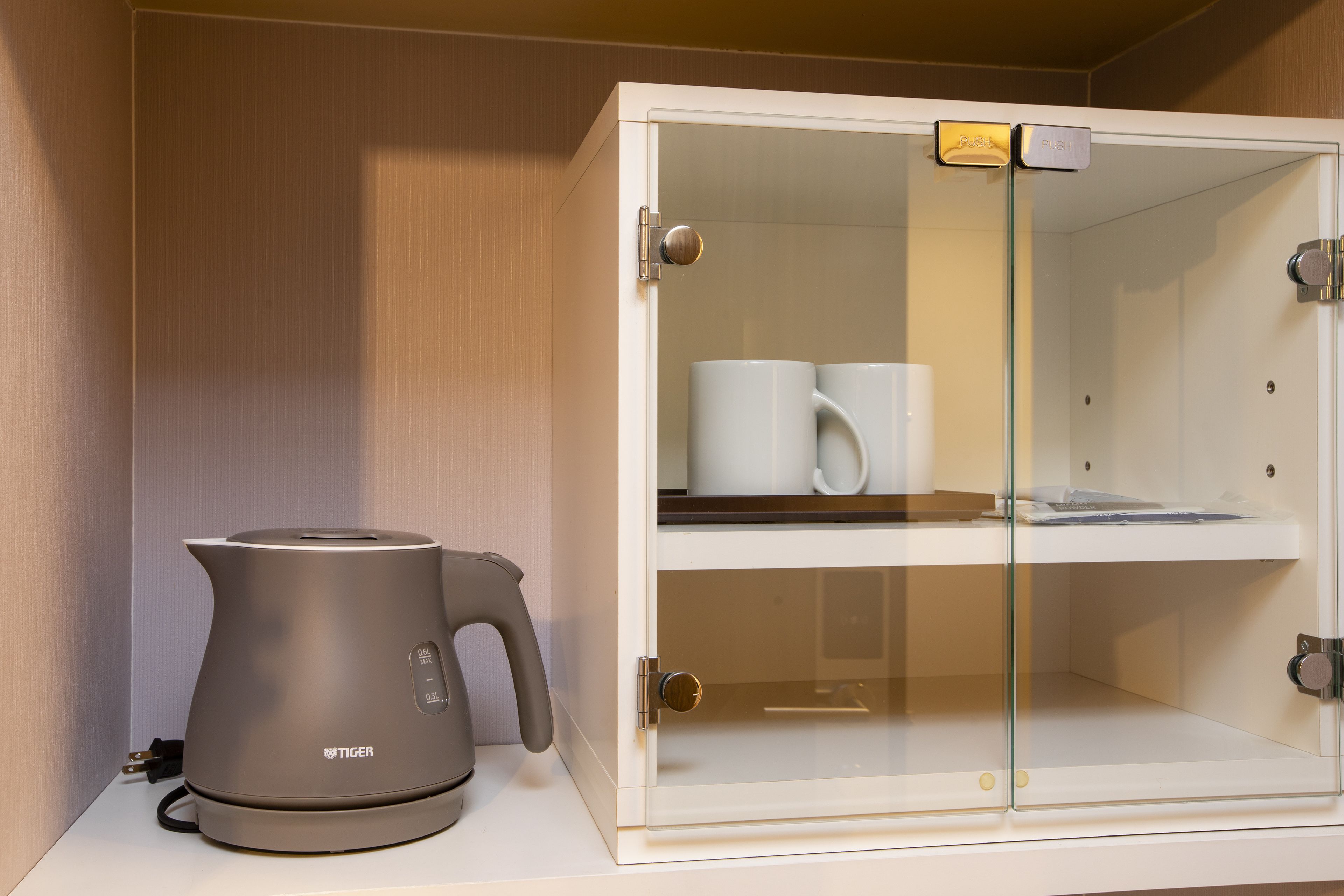 A modern kitchen with a stainless steel kettle and a glass-fronted cabinet containing a coffee maker and cups.