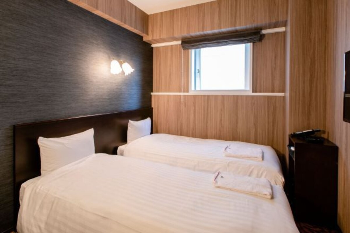A hotel room with two beds, a window, a wall-mounted light fixture, and a dark wooden headboard.