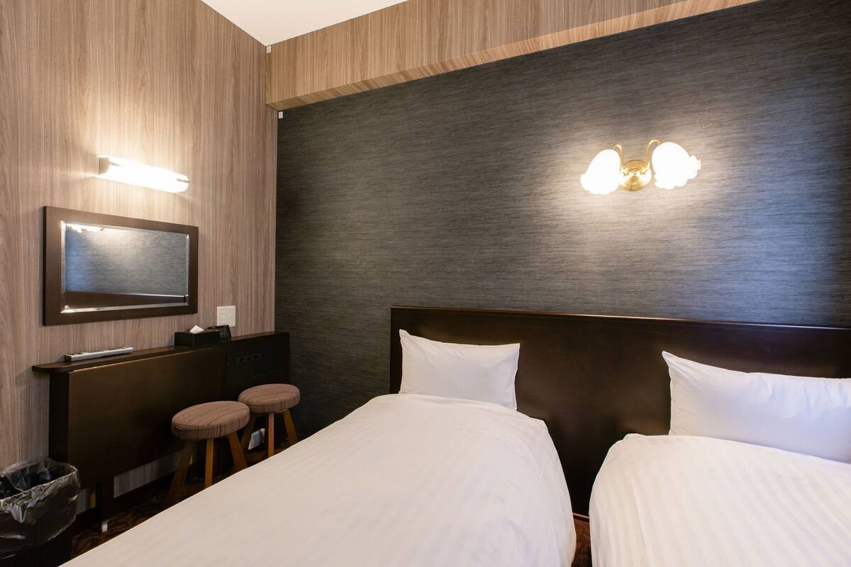 A hotel room with a wooden wall, a dark textured wall, a bed with white bedding, a desk with a computer, and a trash bin.