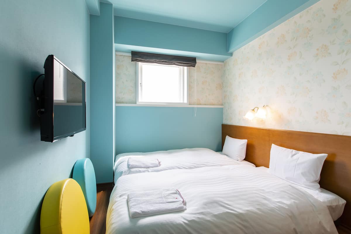 A hotel room with a bed, a television, a window with a curtain, and a wall with floral wallpaper.