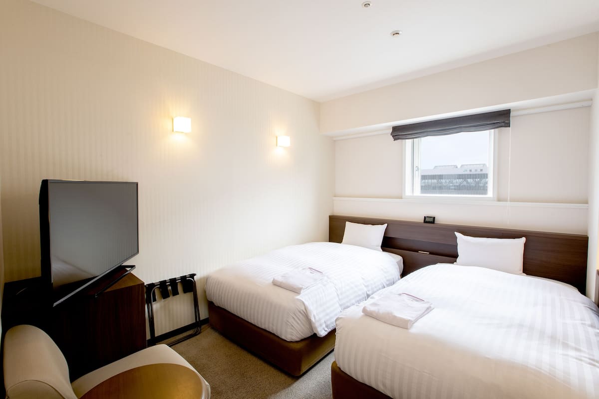 A hotel room with two beds, a TV, a window with a view, and a wall-mounted light.
