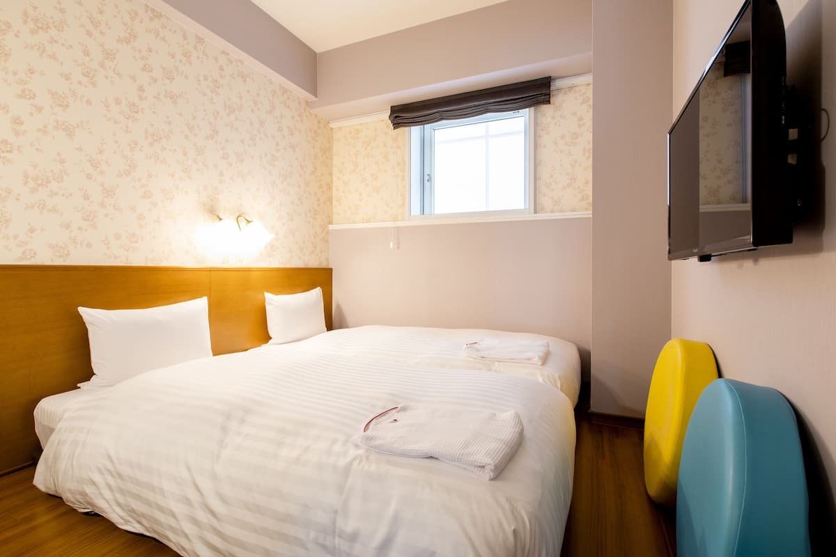 A hotel room with a large bed, two pillows, a bedside table, a flat-screen TV mounted on the wall, and a window with a curtain.