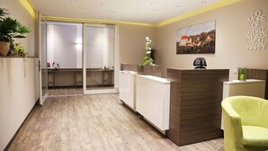 Un área de recepción con suelo de madera, un mostrador, una silla verde, una planta en maceta y un cuadro en la pared.