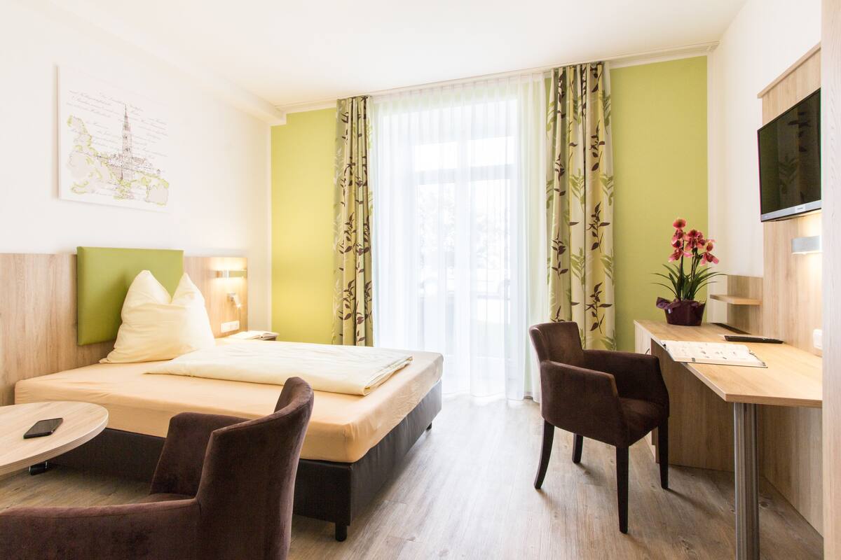 A hotel room with a bed, two chairs, a desk, a TV, and a window with curtains.