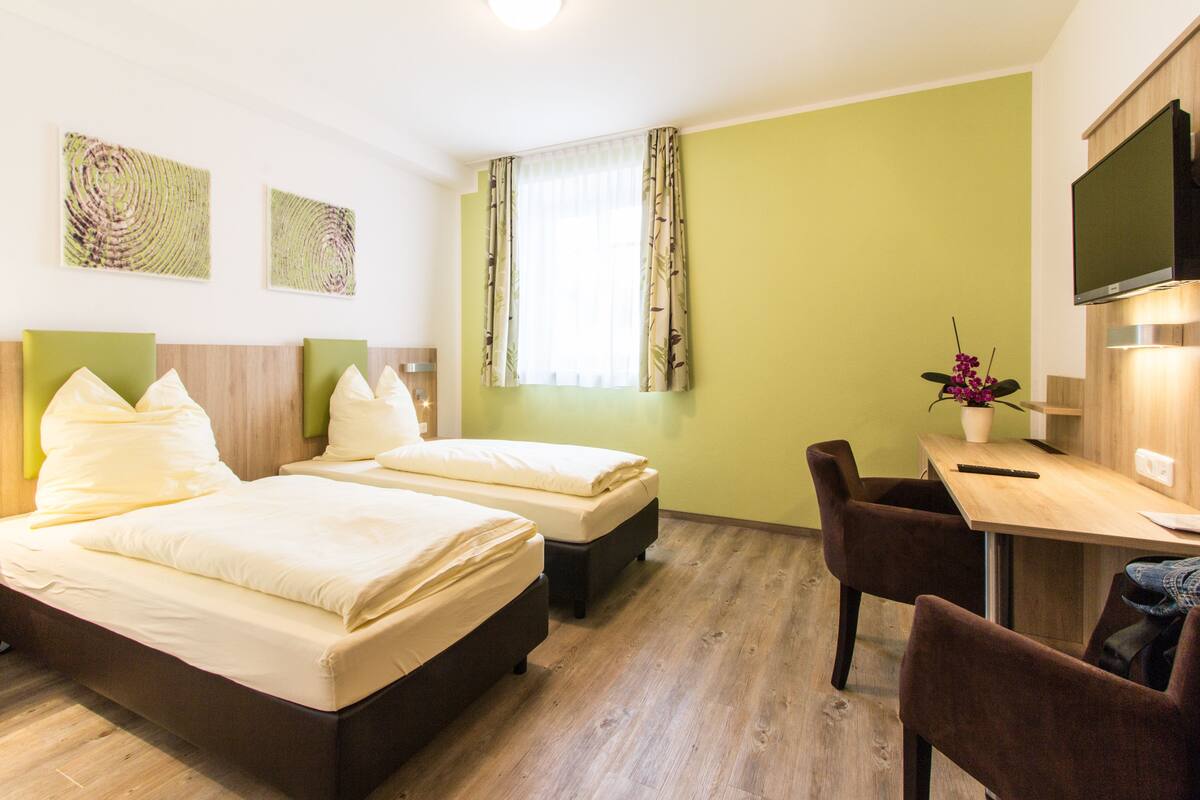 A hotel room with two beds, a desk, a chair, a TV, and artwork on the walls.