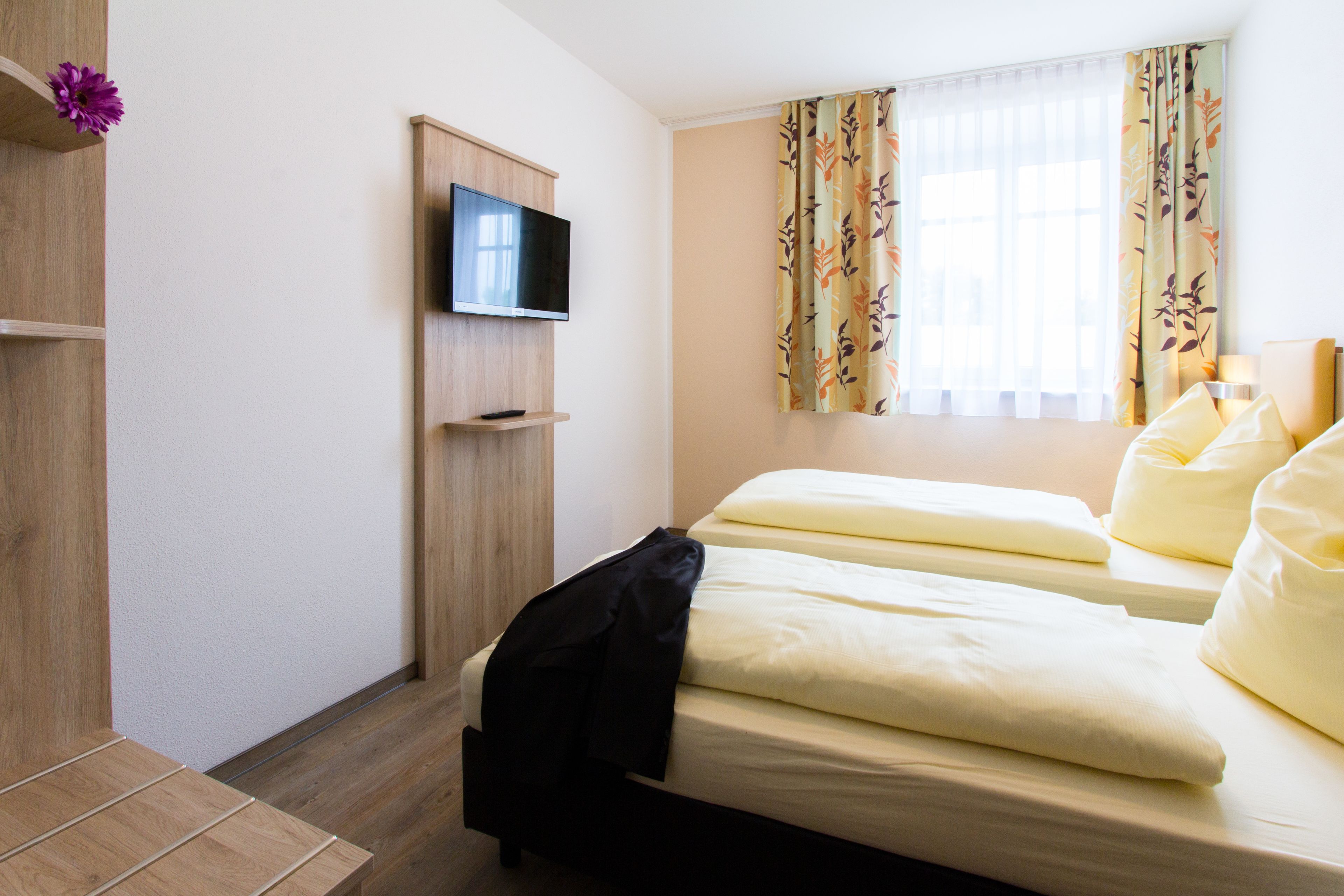 A hotel room with two beds, a TV mounted on the wall, and a window with floral curtains.