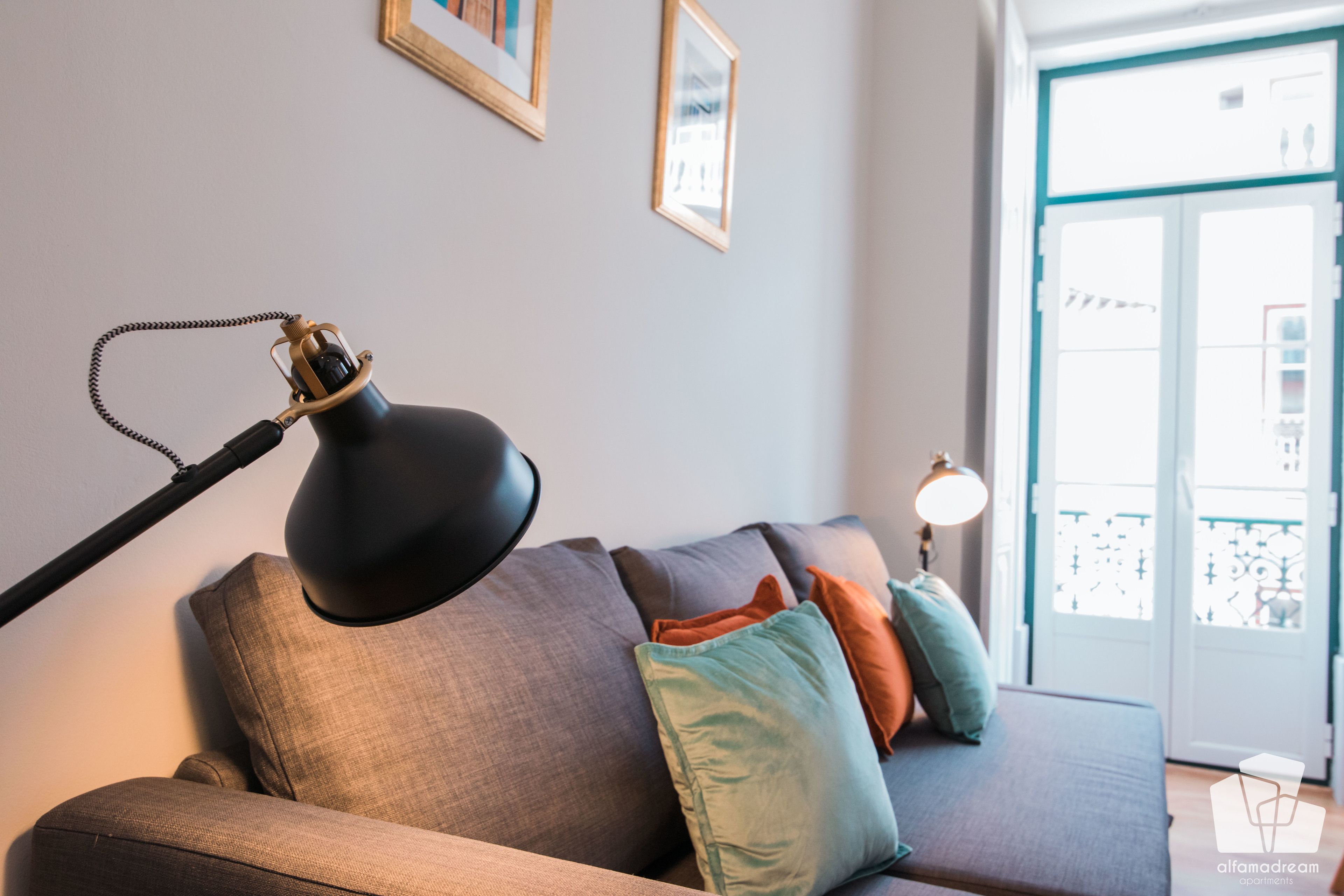 Una sala de estar moderna con un sofá, una lámpara de pie y una lámpara de mesa. Se ven cuadros en la pared y un balcón a través de la ventana.