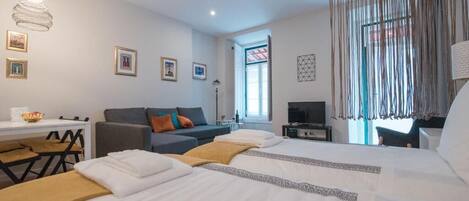 Habitación de hotel con dos camas, un escritorio, una silla, un televisor y un ventanal grande con cortinas.