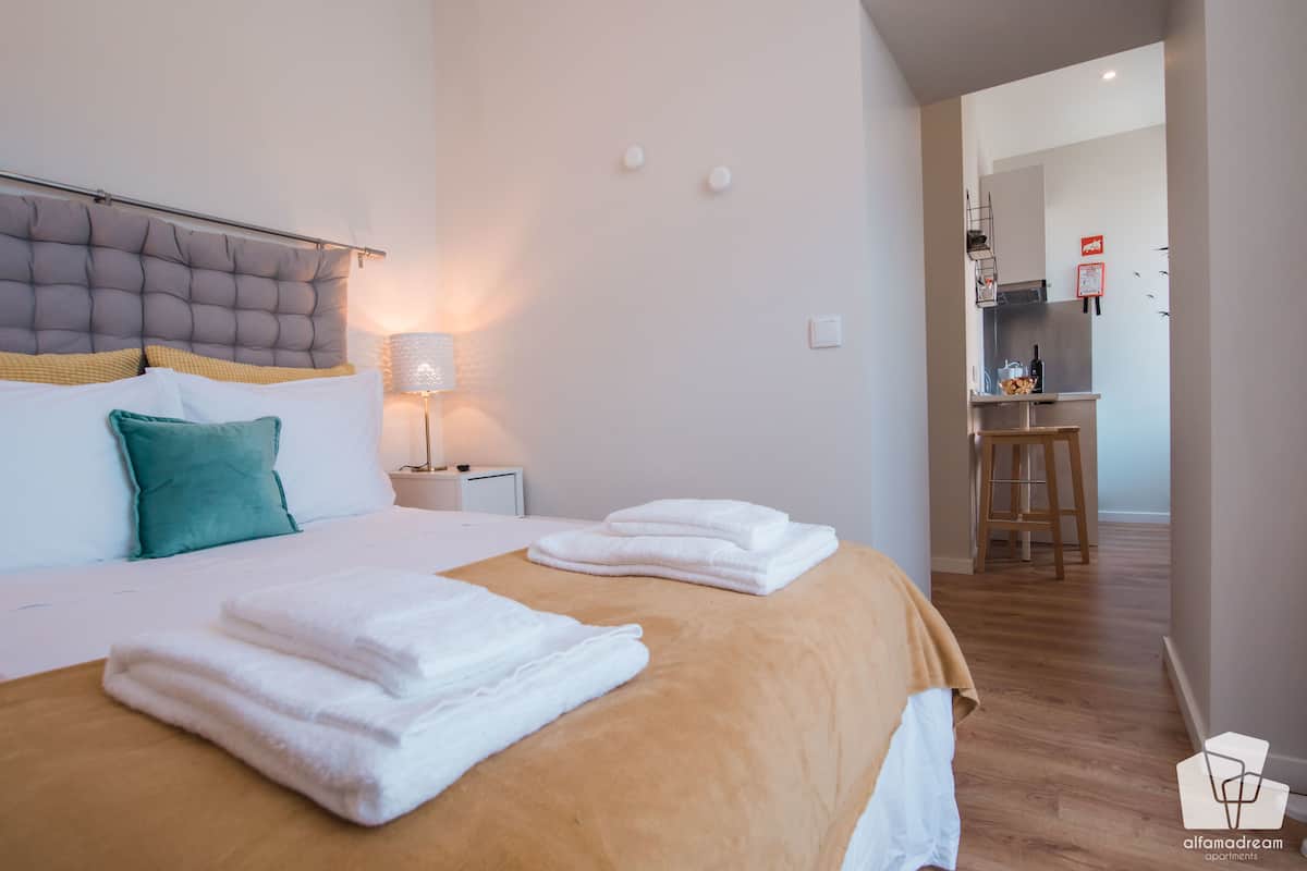 Een hotelkamer met een groot bed, een nachtkastje met een lamp, een kitchenette op de achtergrond en een houten vloer.