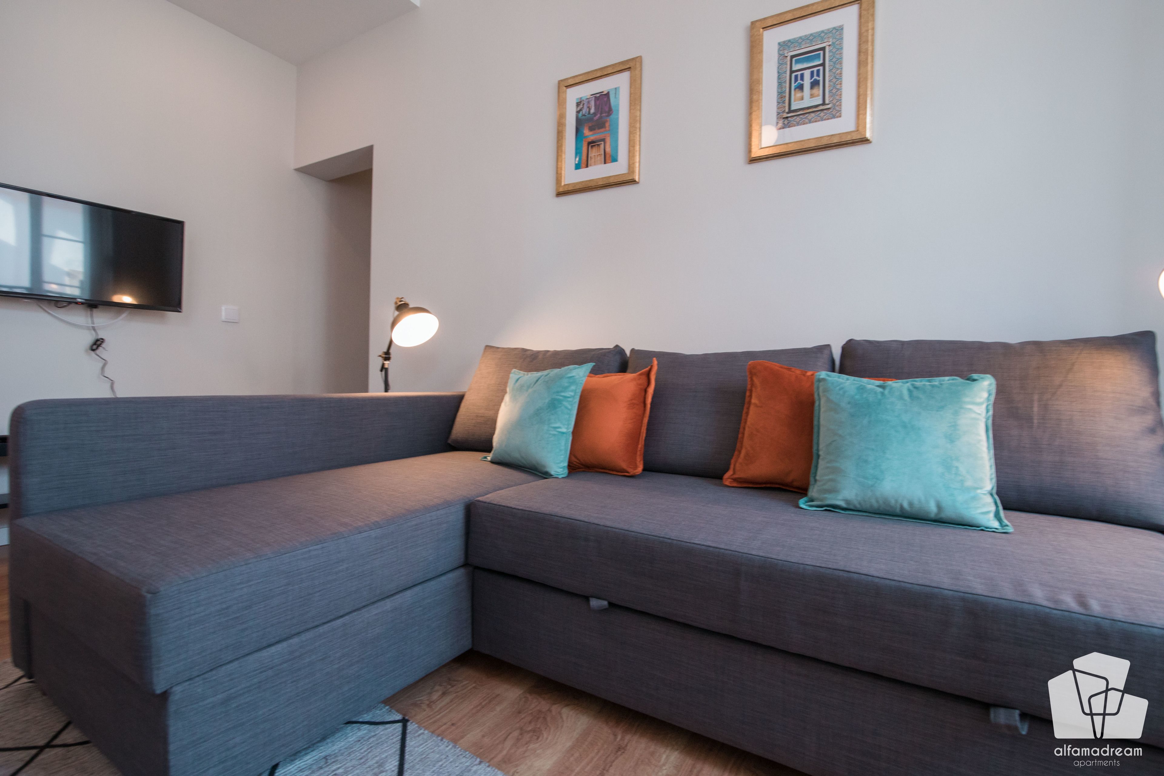 Un salon moderne avec un canapé d’angle gris, un téléviseur à écran plat fixé au mur et deux tableaux encadrés au-dessus du canapé.
