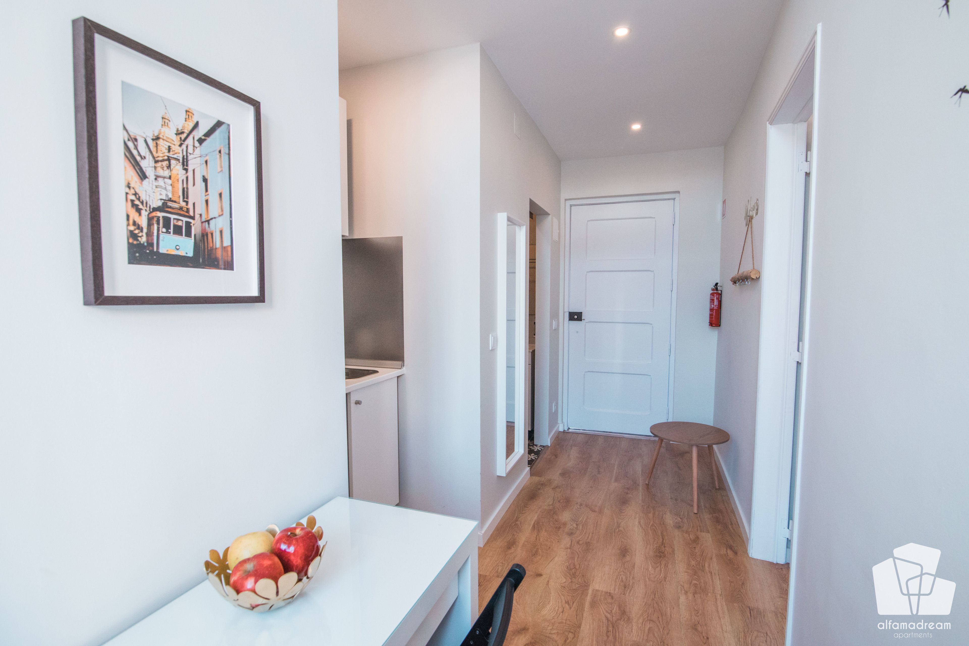 A hallway with a white door, a small table with a bowl of fruit, and a framed picture on the wall.