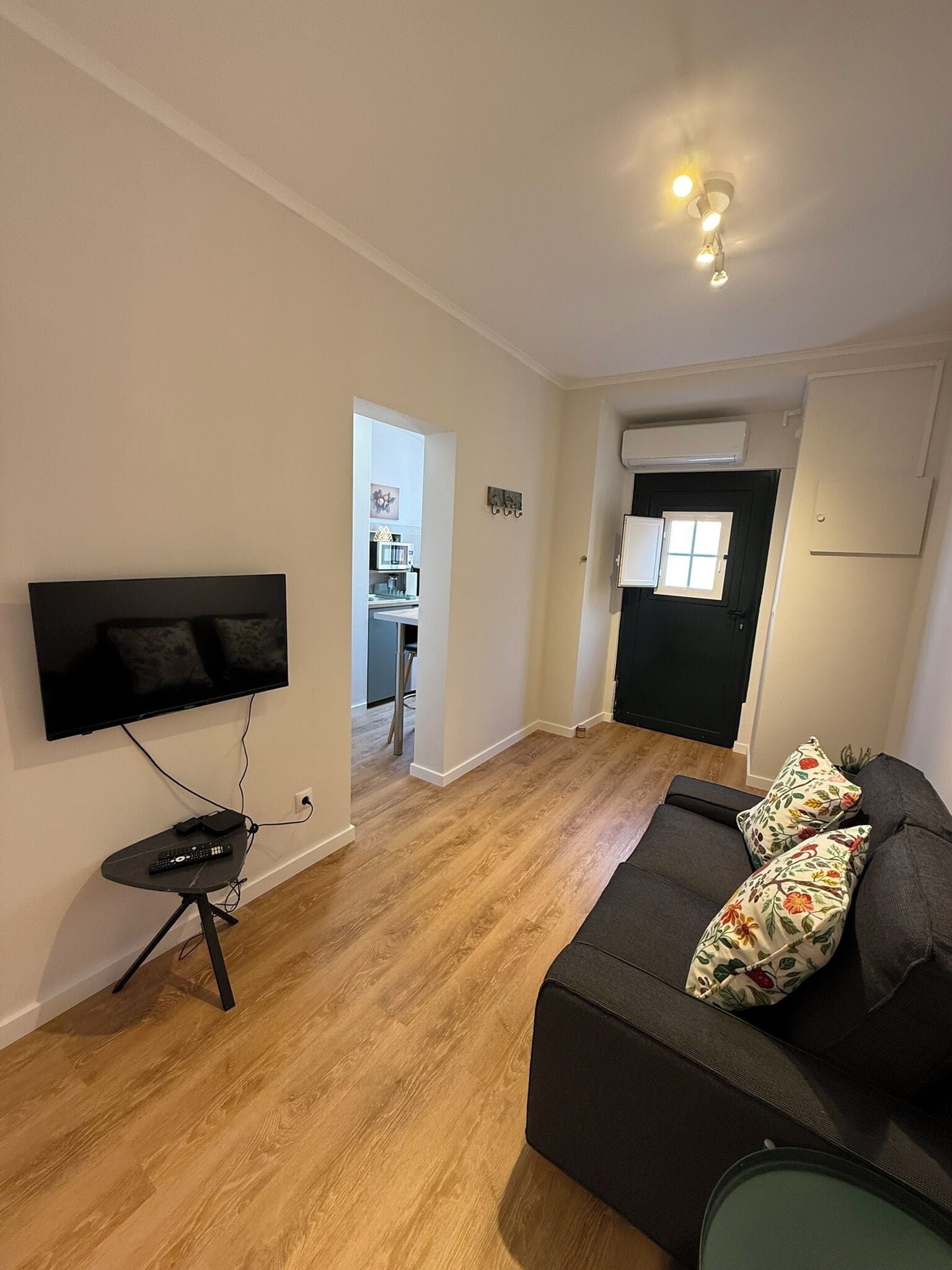 A living room with a flat-screen TV, a small black table with a remote, a dark sofa with floral cushions, and a wooden floor.