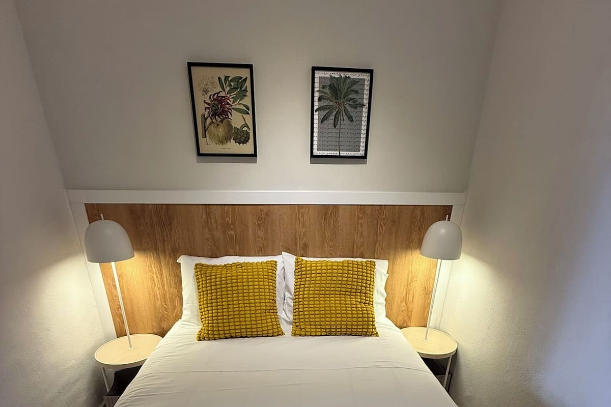 Een keurig opgemaakt bed met witte lakens en twee gele kussens, met aan weerszijden nachtkastjes met bedlampjes.
