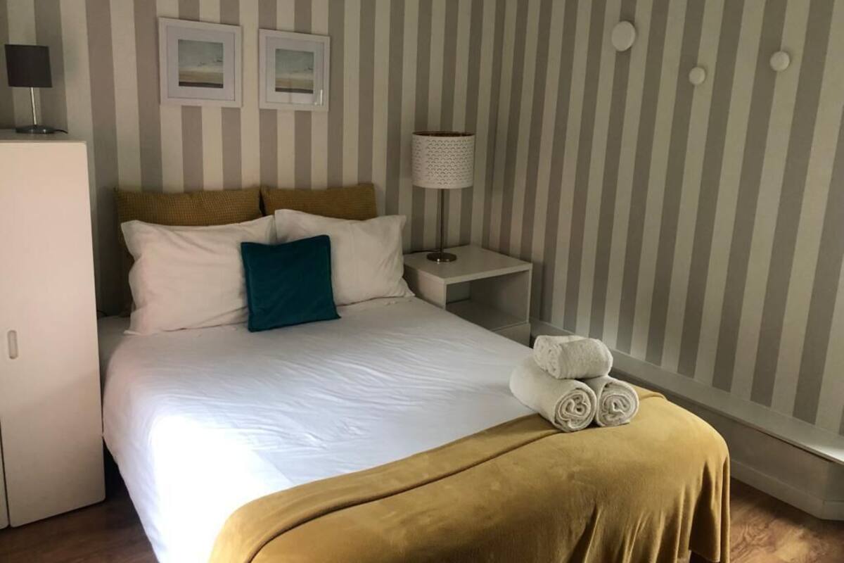 Een net opgemaakt bed met witte en teal kussens, een mosterdgele plaid en opgerolde handdoeken op het bed.