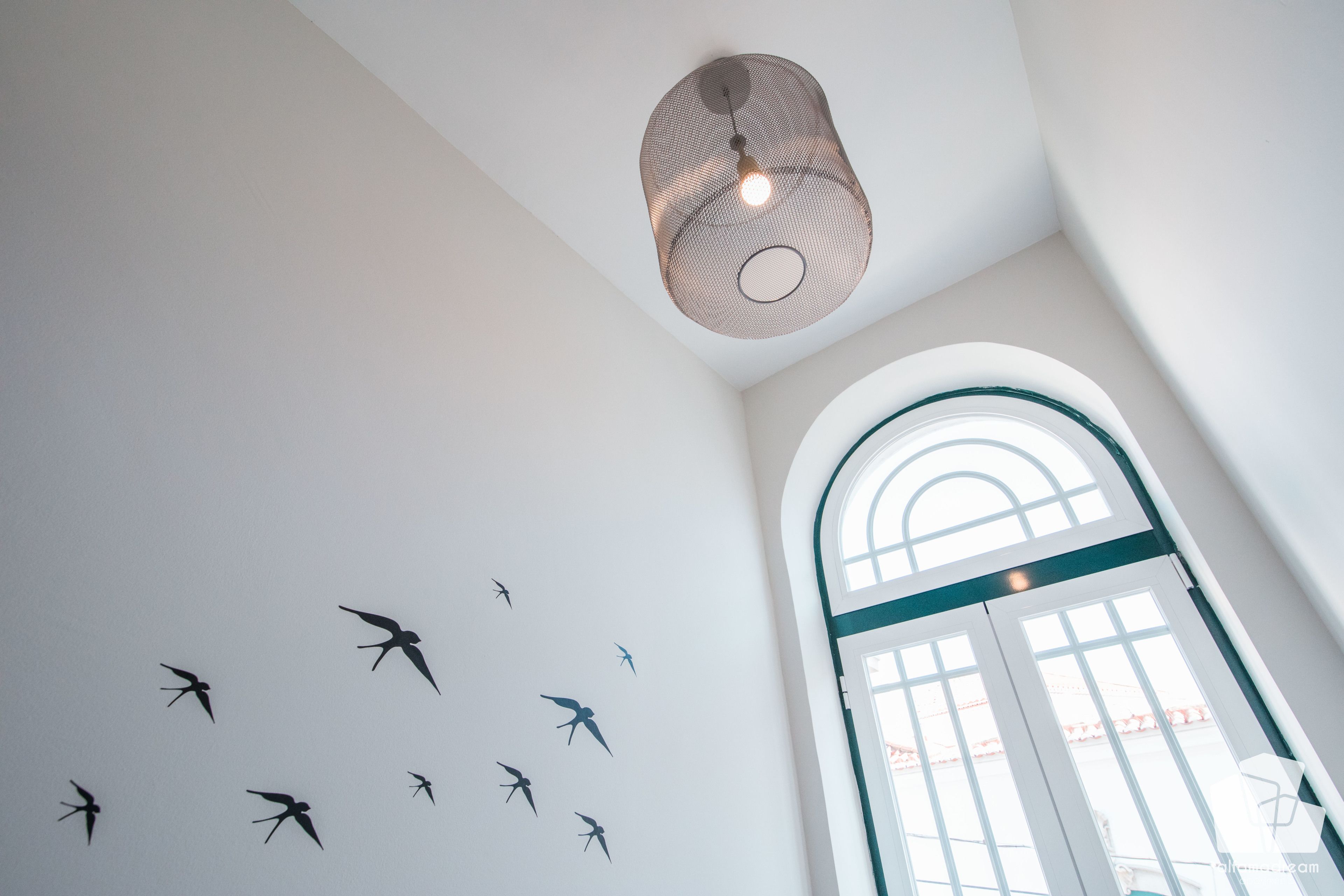 Une pièce avec un plafond haut, un luminaire suspendu et un mur orné de silhouettes d’oiseaux.