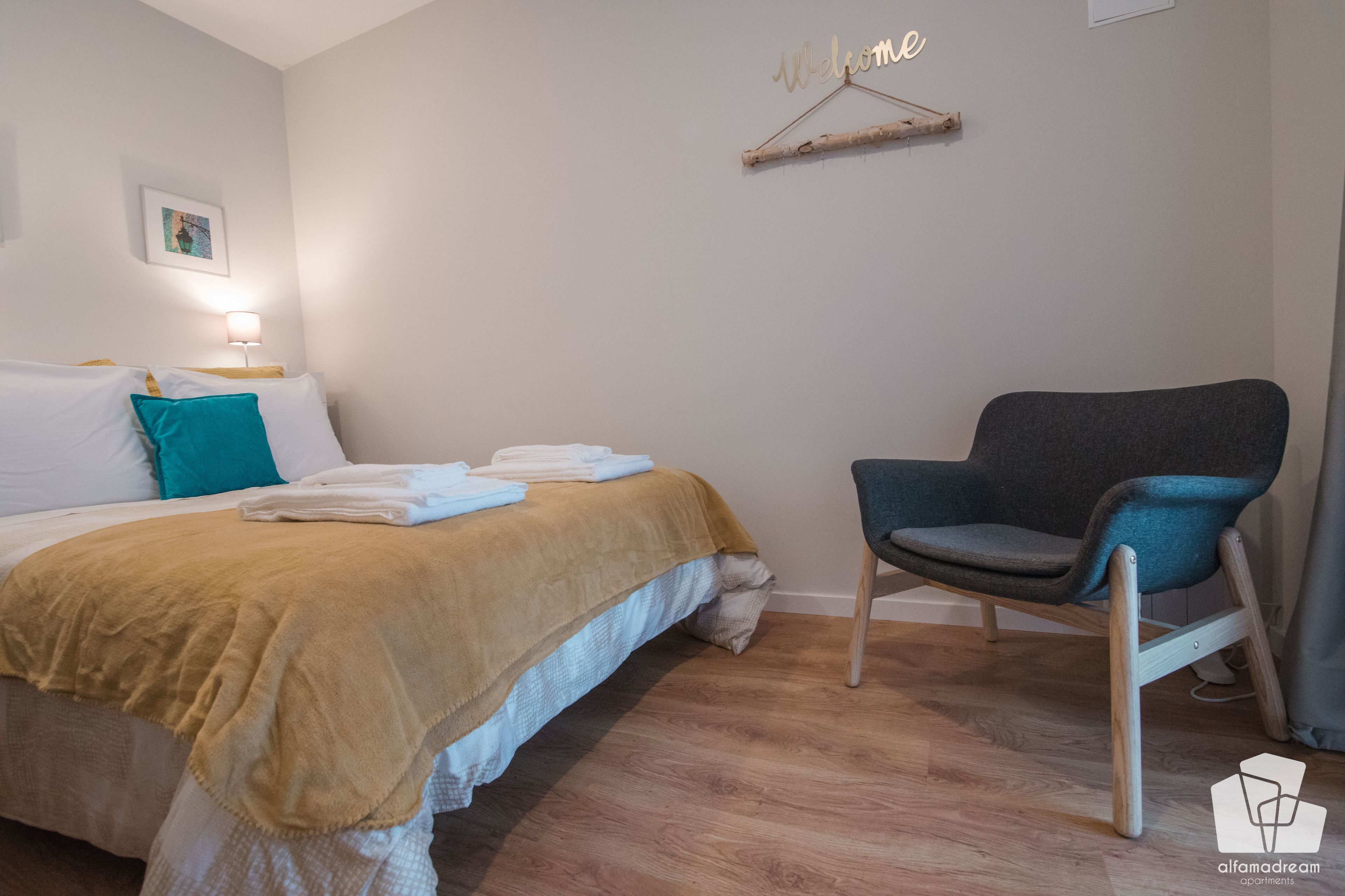 A neatly made bed with a beige blanket, a teal pillow, and folded towels. A dark blue chair is positioned to the right of the bed.