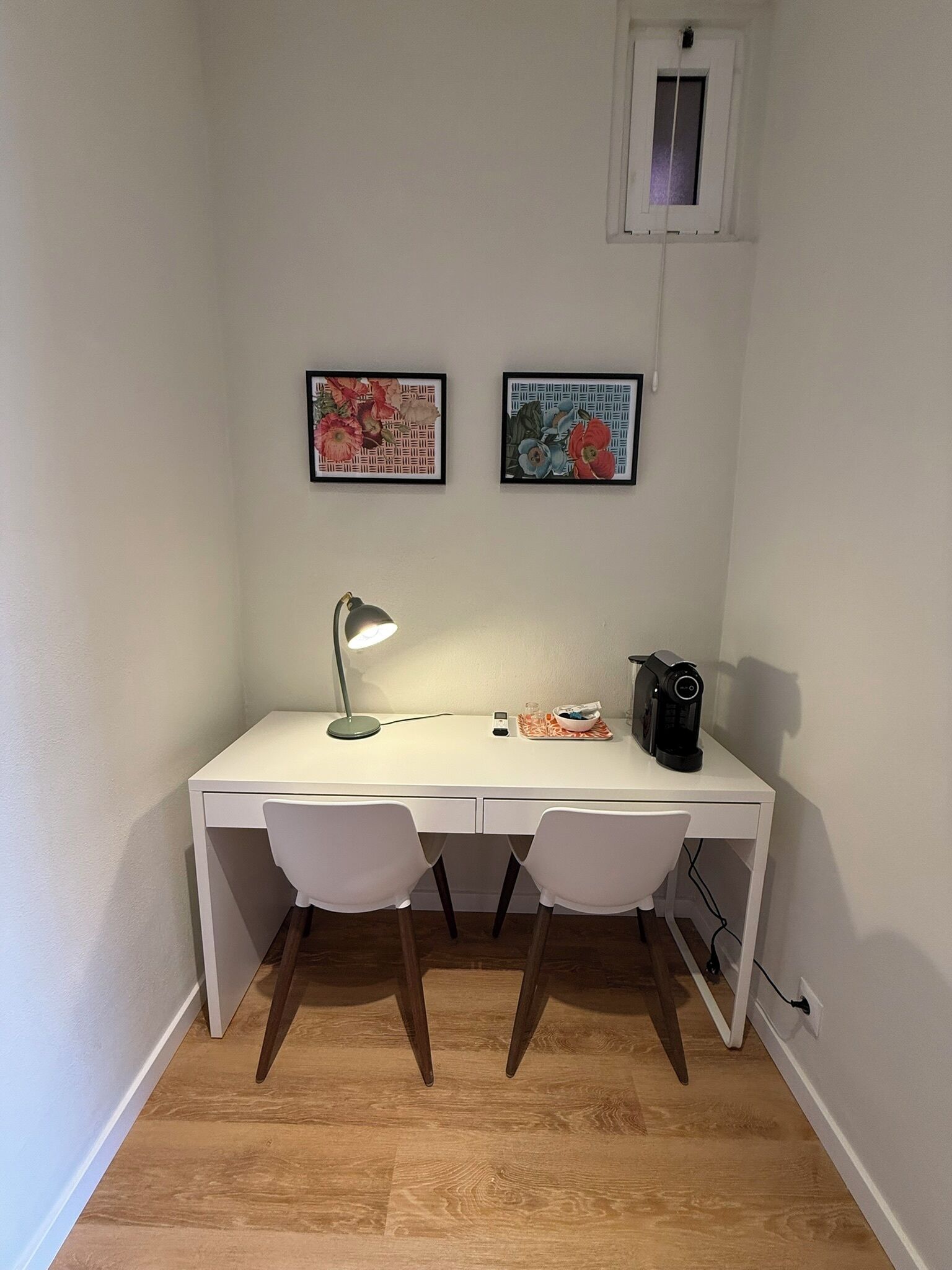 Et hvidt skrivebord med to hvide stole, en skrivebordslampe, en smartphone, en kaffemaskine og to billeder i rammer på væggen.