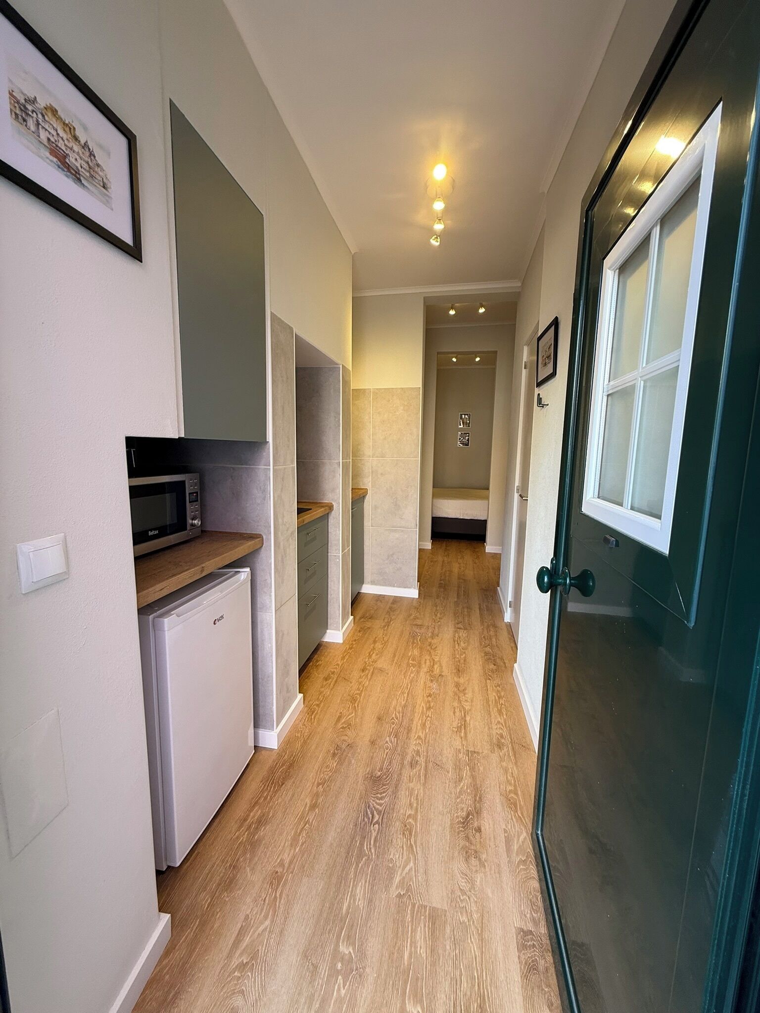Um corredor estreito com piso de madeira, um micro-ondas, uma geladeira e uma porta que dá para outro quarto.