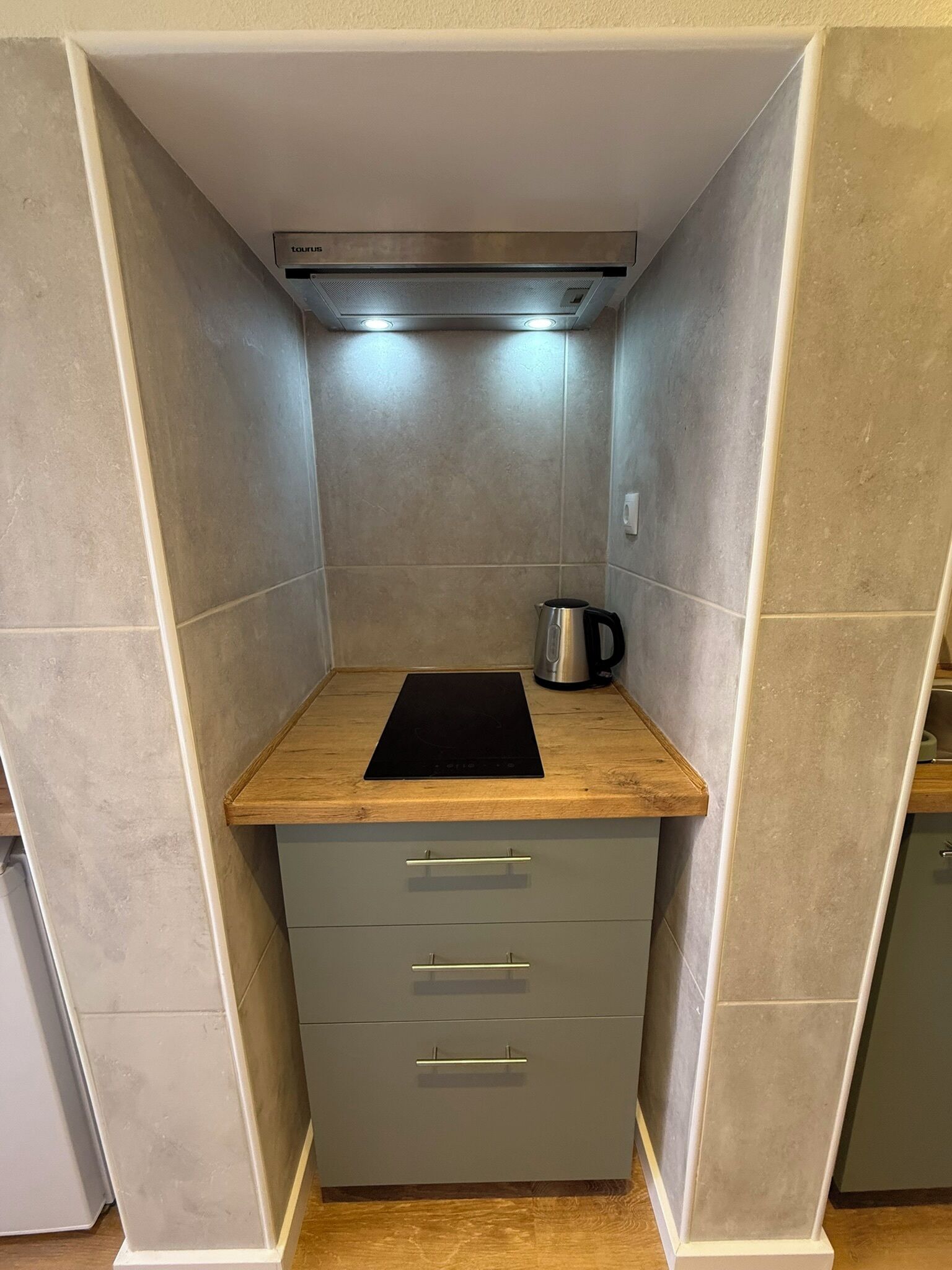 Een compacte kitchenette met een ingebouwde kookplaat, een waterkoker en een klein kastje.
