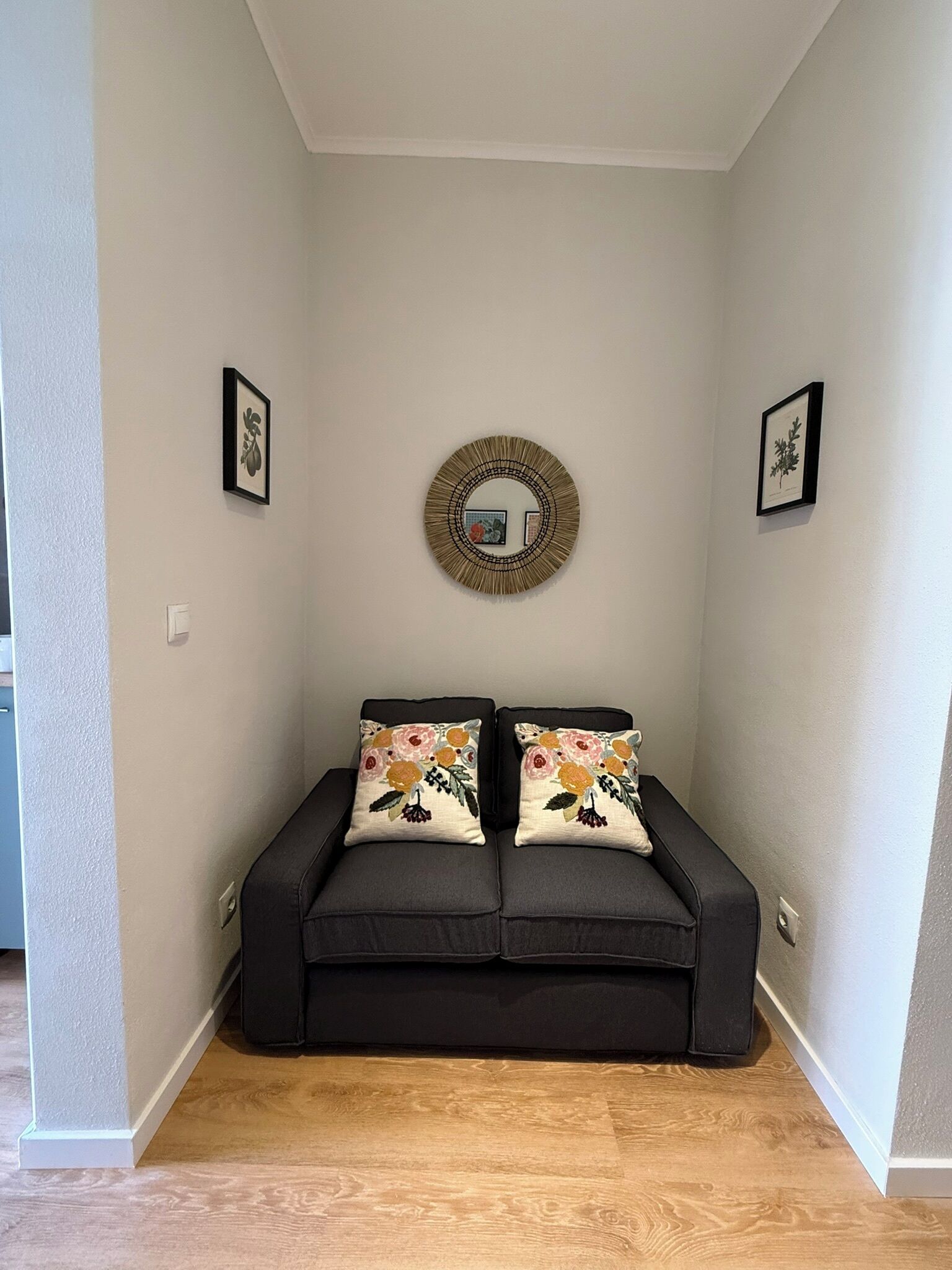 A small, cozy room with a dark-colored sofa, two decorative pillows, a round mirror, and framed artwork on the walls.