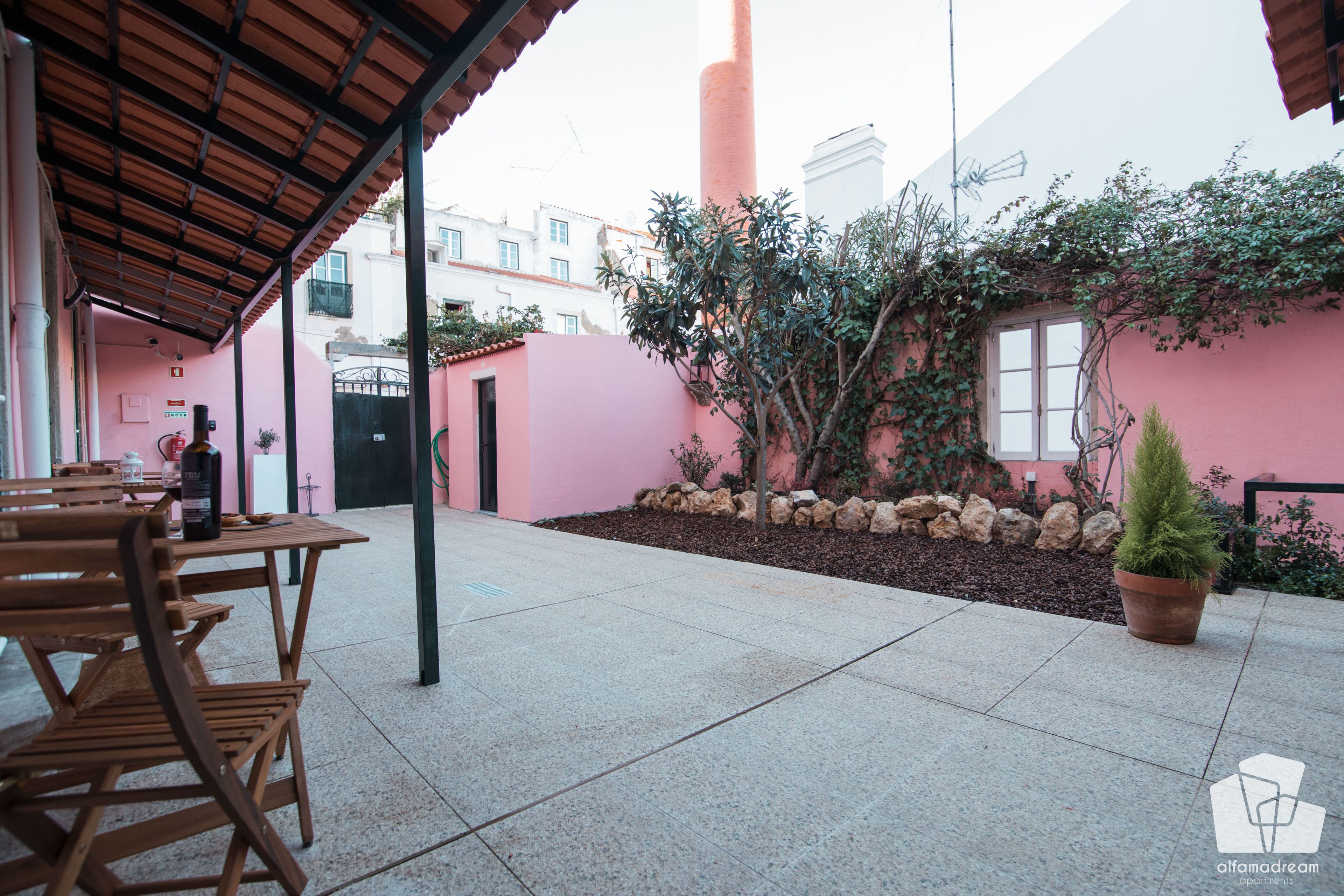 Une cour avec un sol carrelé, une table et des chaises en bois, une plante en pot et un mur rose orné de plantes grimpantes.