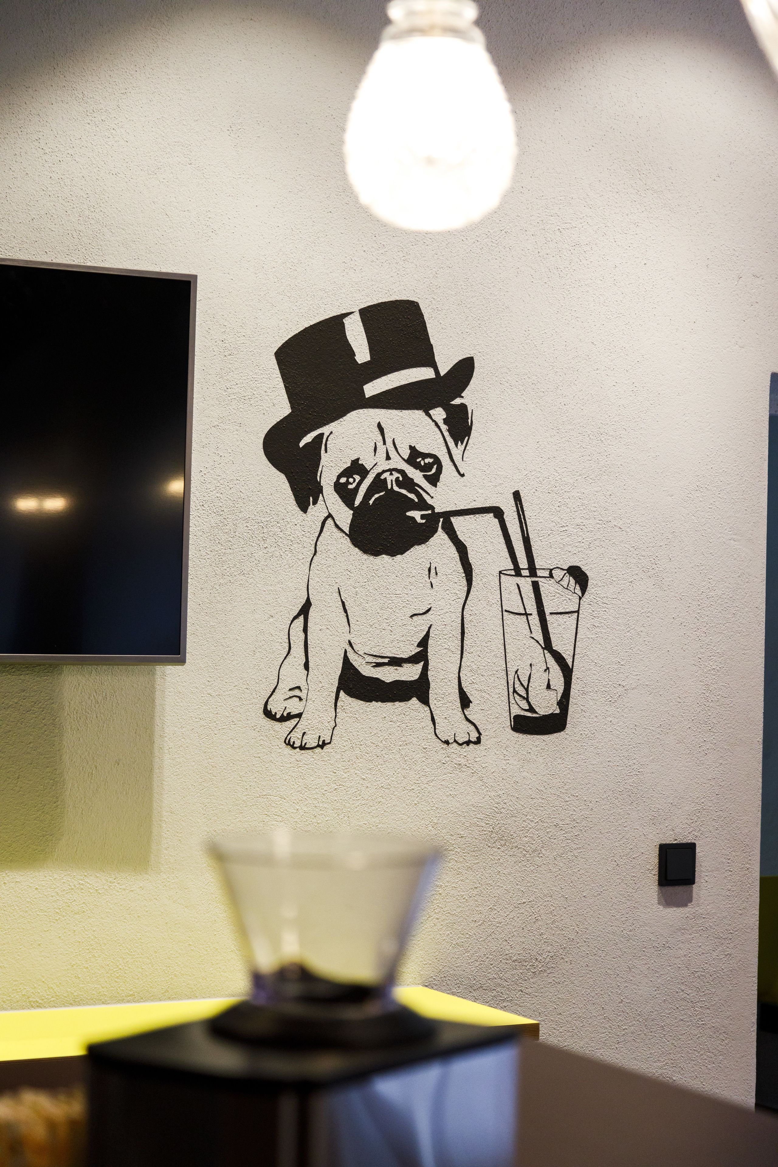 Un mur orné d’une fresque en noir et blanc représentant un chien portant un chapeau haut de forme et tenant un verre à la main.