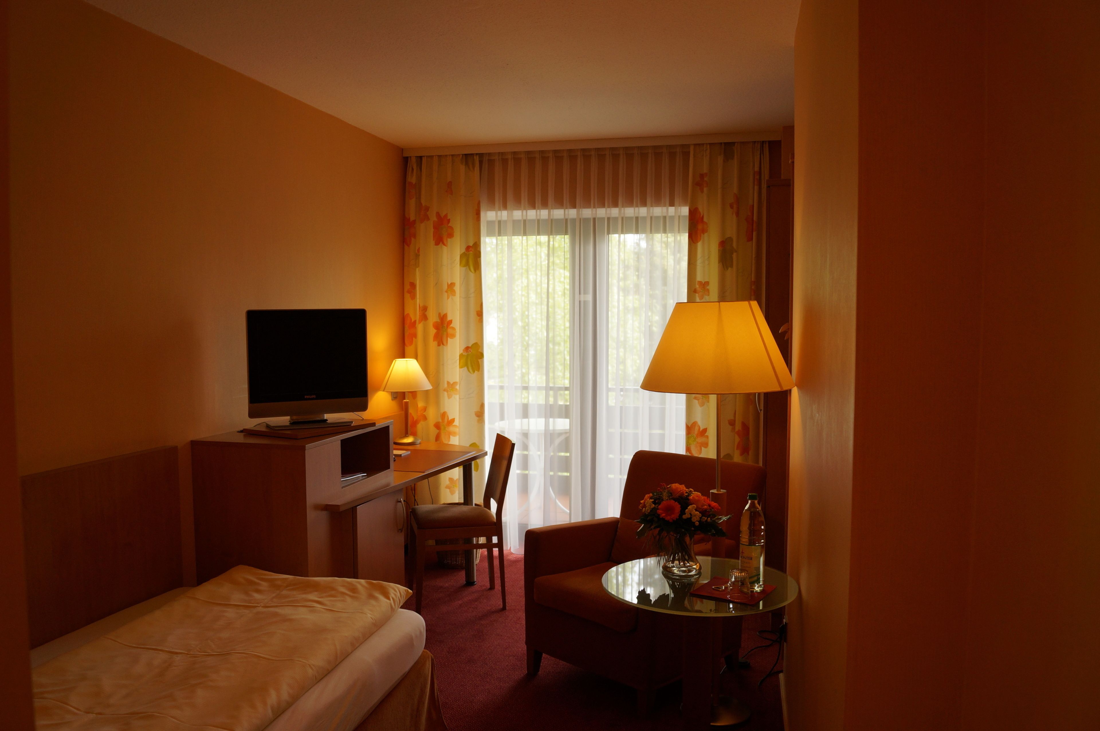 Ein Hotelzimmer mit einem Bett, einem Schreibtisch mit Fernseher, einem Sessel, einem Tisch mit Lampe und Blumen sowie einem Fenster mit Vorhängen.