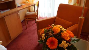 A hotel room with a wooden desk, a chair, a bedside table with a vase of flowers, and a window with curtains.