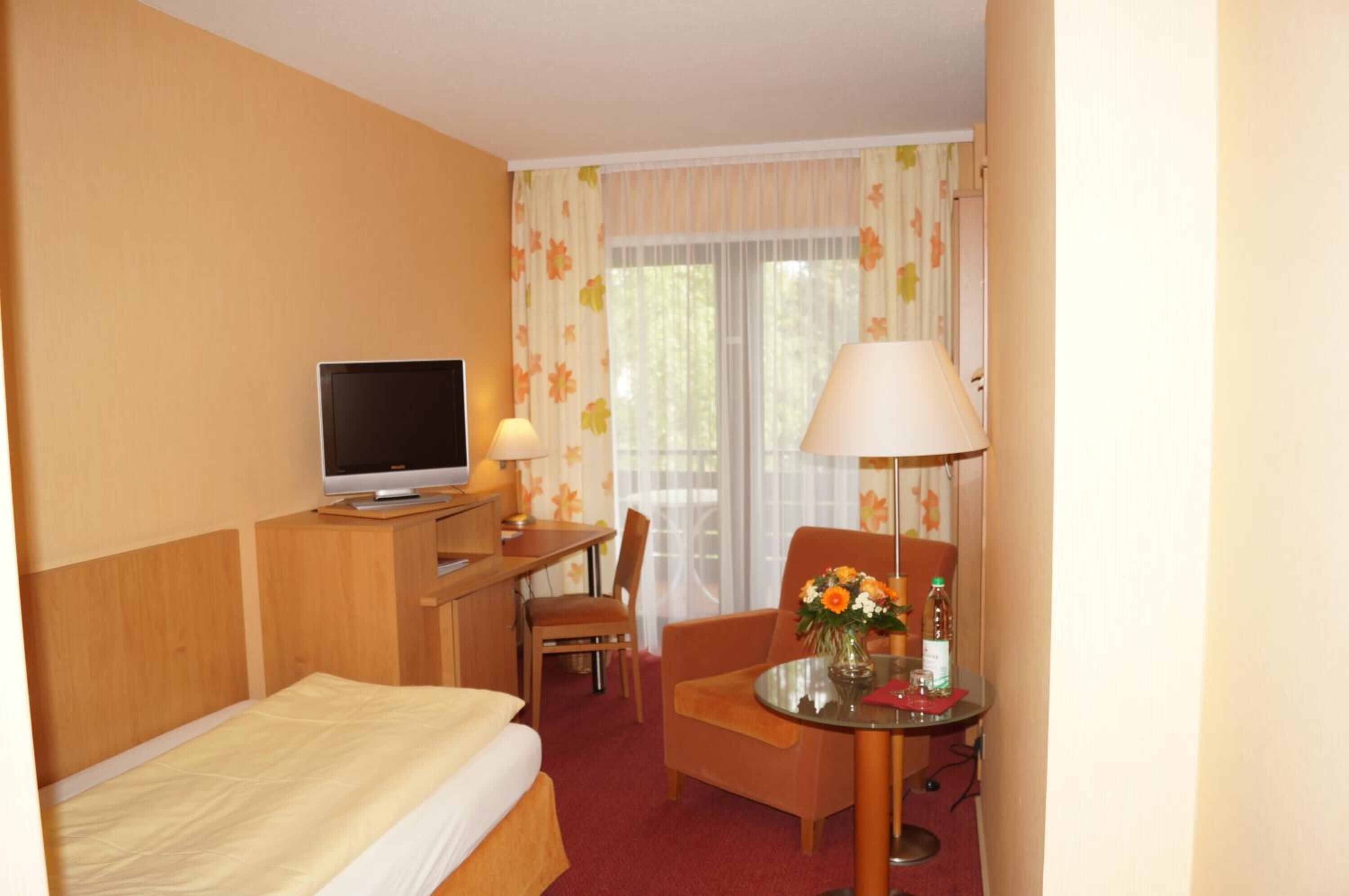 A hotel room with a bed, a desk with a computer, a chair, a table with a vase of flowers, and a window with curtains.