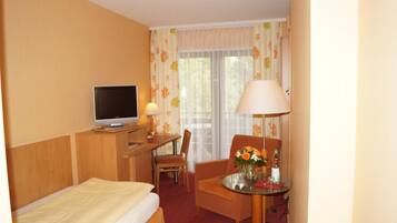 A hotel room with a bed, a desk with a computer, a chair, a table with a vase of flowers, and a window with curtains.